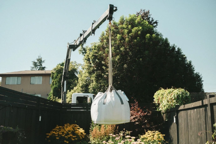 A large white bulk bag of soil being hoisted by a delivery crane over a backyard garden.