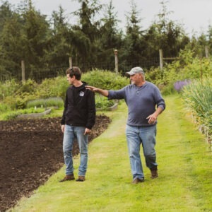 Two men walking and talking together in a vibrant garden with one man gesturing to the lush surroundings, demonstrating or explaining something to the other.