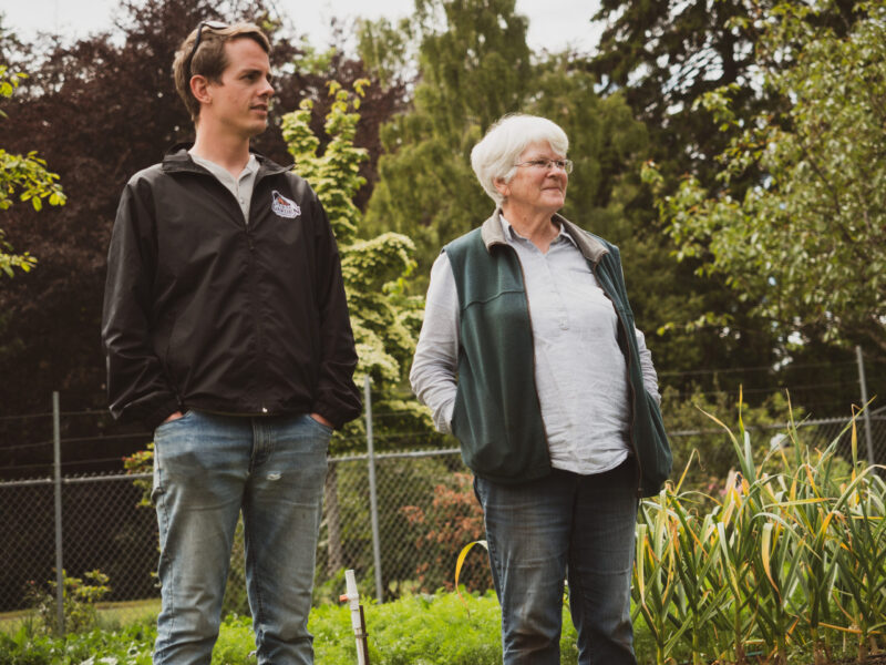 A young man and an elderly woman stand together in a garden, looking off to the side. both are dressed casually, surrounded by greenery and plants.