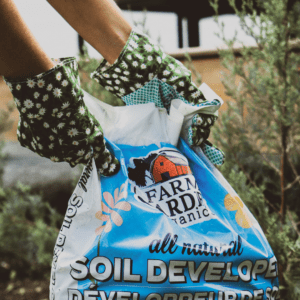 Person wearing patterned gardening gloves holds a large bag of organic soil developer in a lush garden setting.